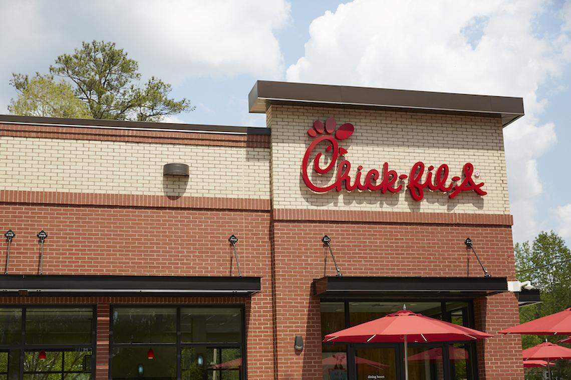 The exterior of a Chick-fil-A restaurant features a classic red and tan brick facade under a bright, partly cloudy sky. The iconic red script logo is mounted prominently above the entrance, which is shaded by vibrant red patio umbrellas.