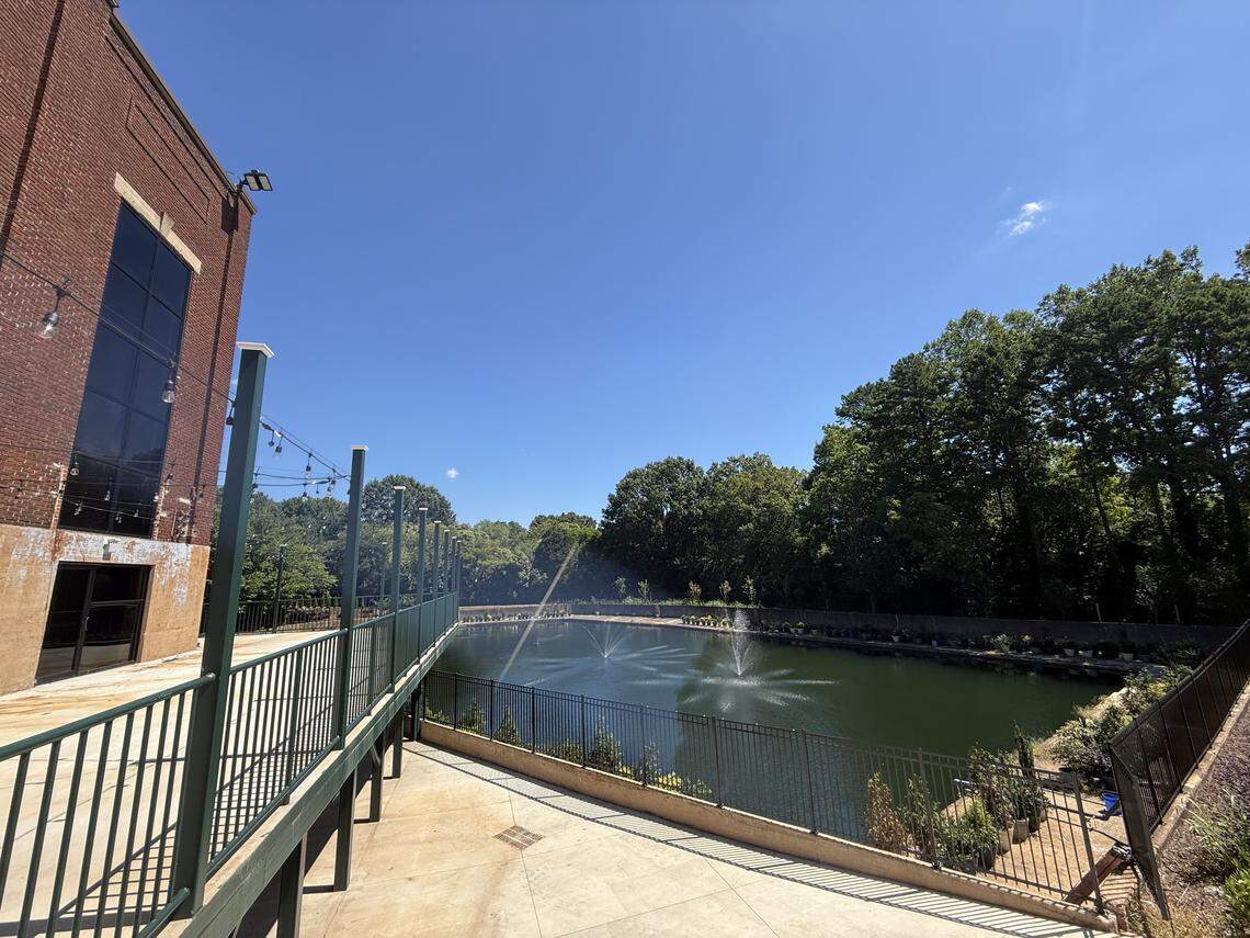 A high-angle view of a serene pond with two fountains, located next to a large brick building. The pond is surrounded by a black metal fence and lush green trees under a clear blue sky.