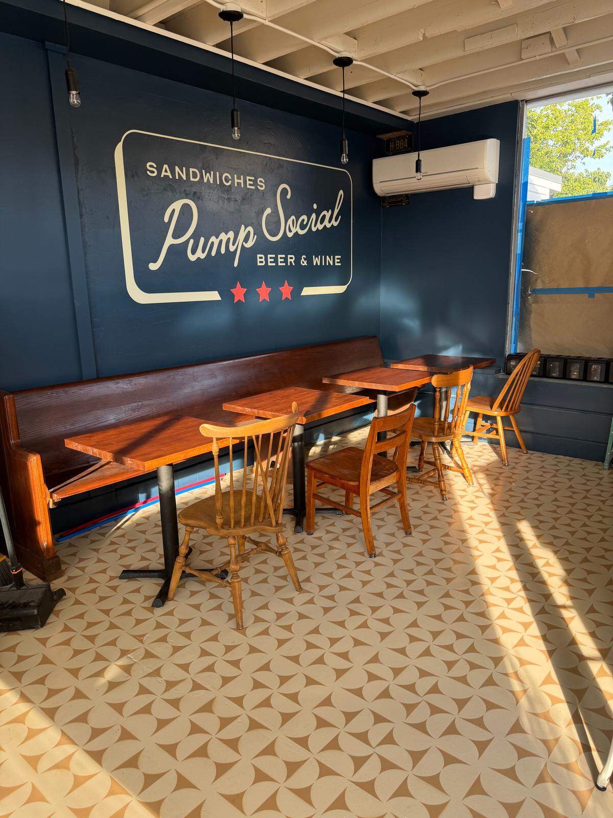 A sunlit corner of a cafe’s interior shows a long bench seat with three small wooden tables and chairs. The wall behind the seating is painted a dark blue, with a large white sign that reads “Sandwiches Pump Social Beer & Wine.”