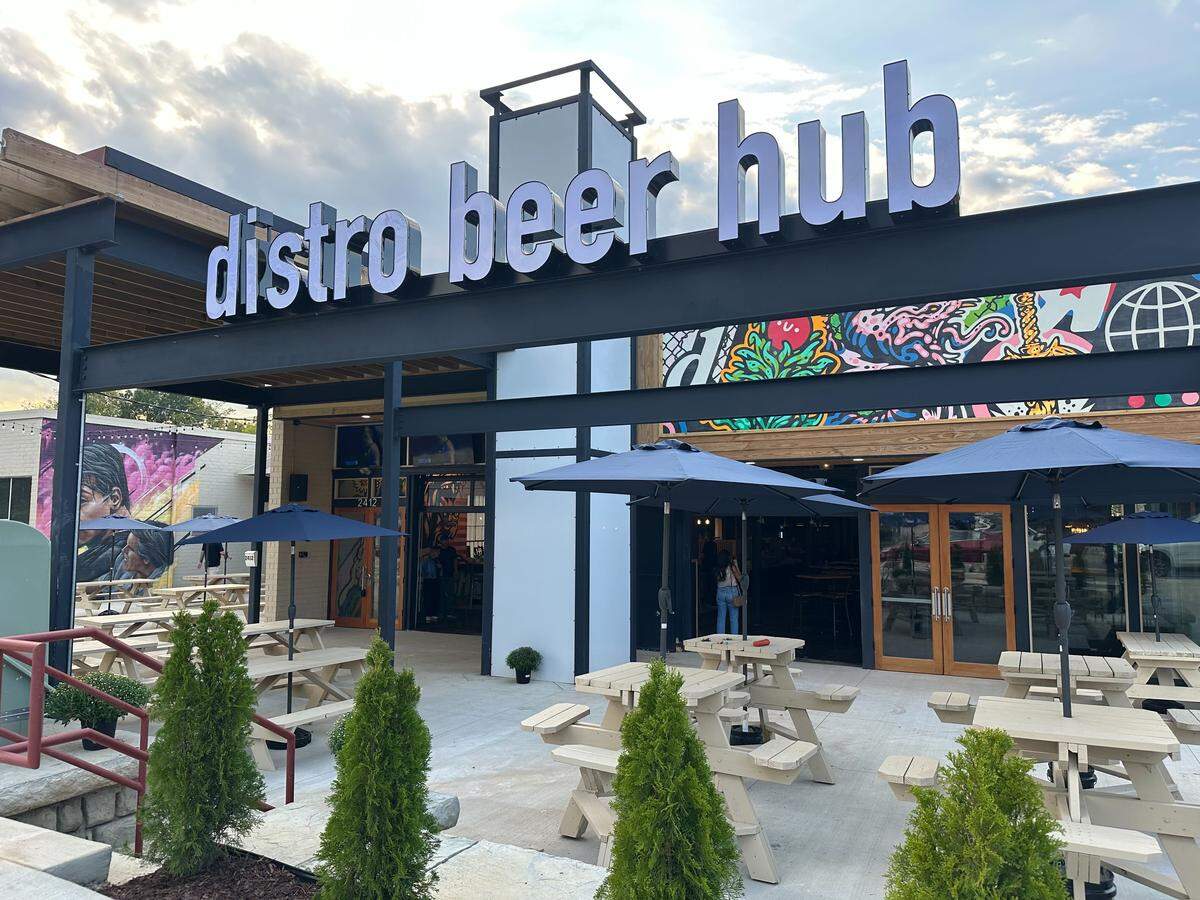 A large, modern building with a sign that reads “distro beer hub” in big, white letters. The building has a patio area with several picnic tables and blue umbrellas.
