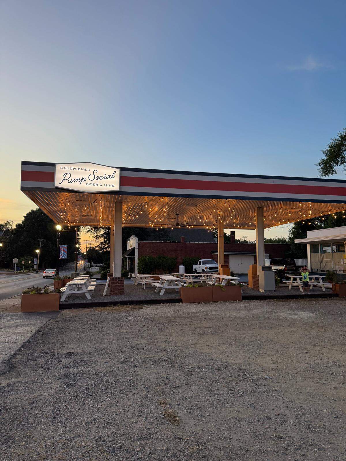A former gas station at dusk has been converted into a restaurant called “Pump Social.” The covered area where gas pumps used to be is now filled with white picnic tables under a canopy of string lights.