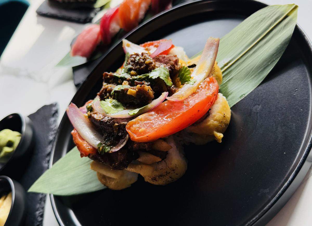 A plate of Lomo Saltado sushi is the focus of a long, white table. The plate holds a large roll topped with steak, red onion, and tomato, with other sushi plates blurred in the background.