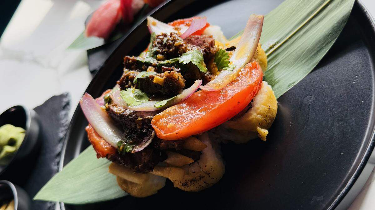 A plate of Lomo Saltado sushi is the focus of a long, white table. The plate holds a large roll topped with steak, red onion, and tomato, with other sushi plates blurred in the background.