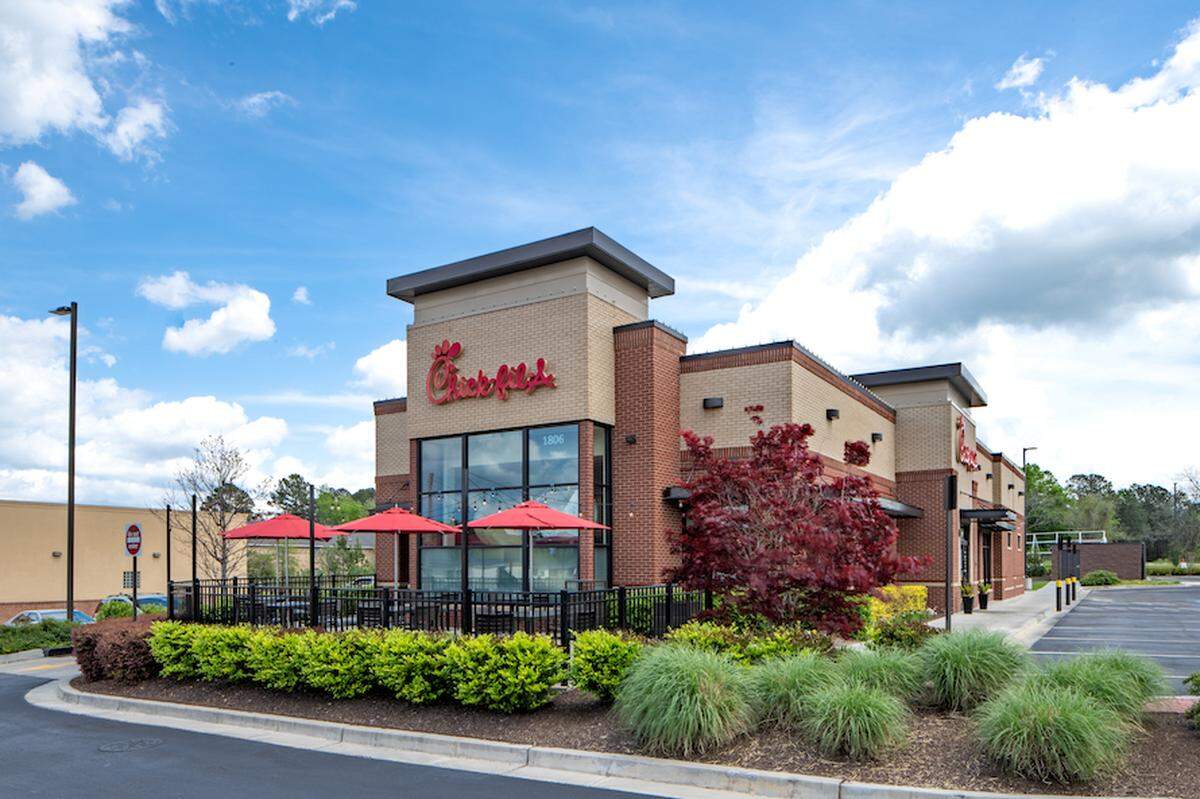 A Chick-fil-A restaurant building with a drive-thru and an outdoor patio with red umbrellas is shown on a partly cloudy day. The building is made of light-colored brick with dark accents and has well-maintained landscaping in the foreground.