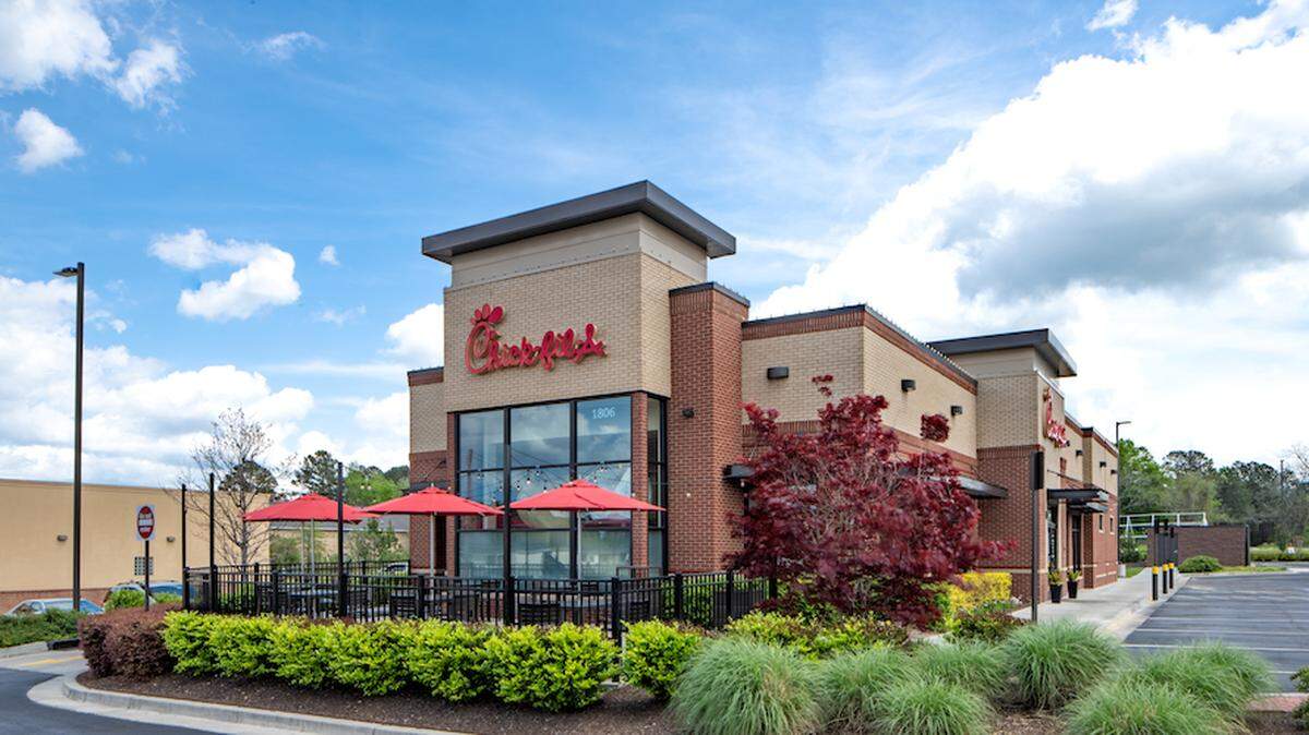 A Chick-fil-A restaurant building with a drive-thru and an outdoor patio with red umbrellas is shown on a partly cloudy day. The building is made of light-colored brick with dark accents and has well-maintained landscaping in the foreground.