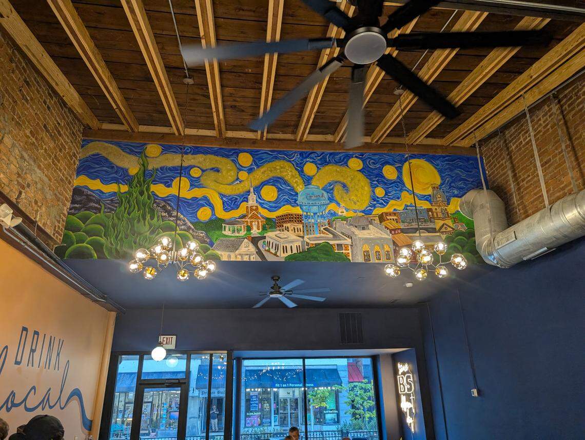 A mural painted in the style of Van Gogh’s “The Starry Night” depicts a small town. The mural is painted on a dark blue wall above a large window in a coffee shop with exposed brick and a wooden beam ceiling.