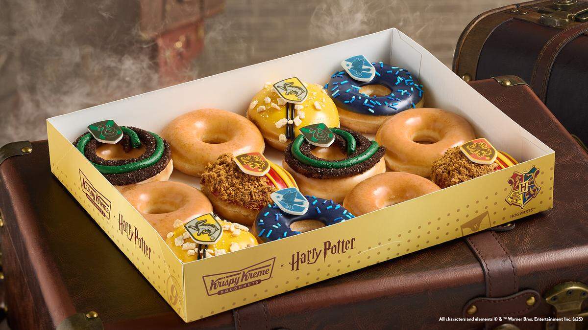 A dozen Harry Potter-themed Krispy Kreme Doughnuts are displayed in an open box on top of a vintage-style trunk. The box contains plain glazed doughnuts and doughnuts decorated with the crests of the four Hogwarts houses: Gryffindor, Ravenclaw, Hufflepuff, and Slytherin.