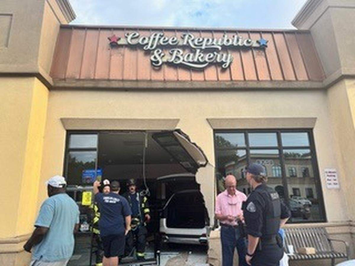 A dark SUV is shown crashed into the front window of a building that reads “Coffee Republic & Bakery.” A group of first responders and onlookers stands outside, assessing the damage.