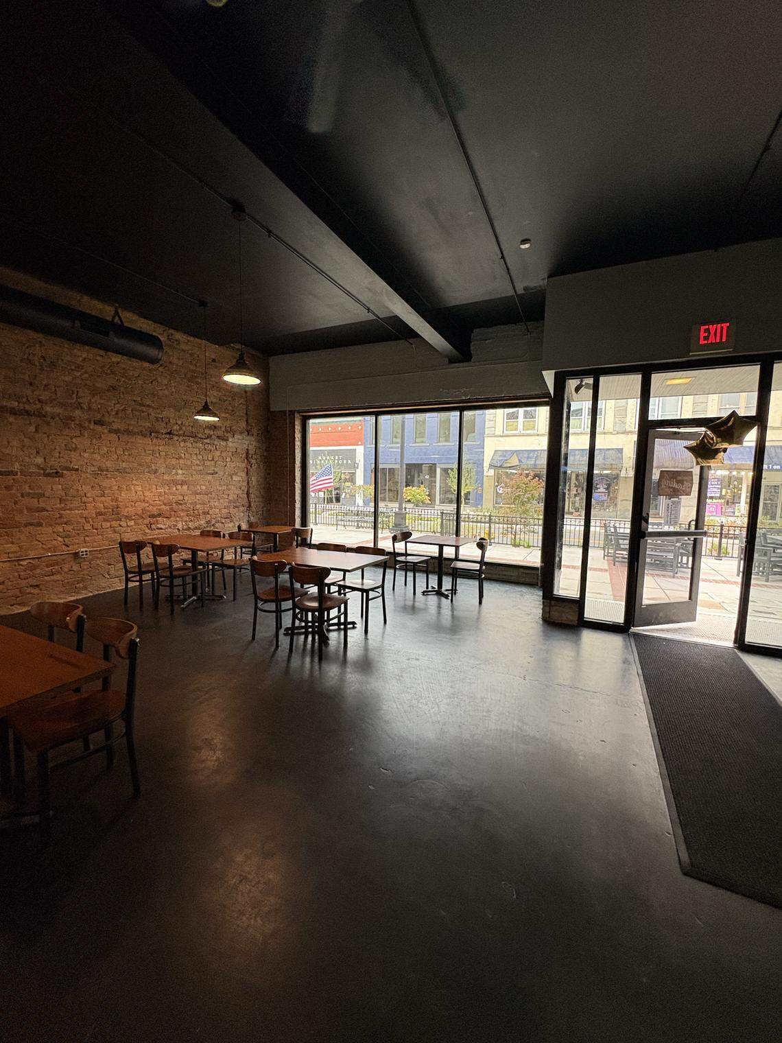 A photo shows the empty interior of a small restaurant or cafe with tables and chairs. The space has a high black ceiling, exposed brick walls, and a large glass facade that looks out onto a street.