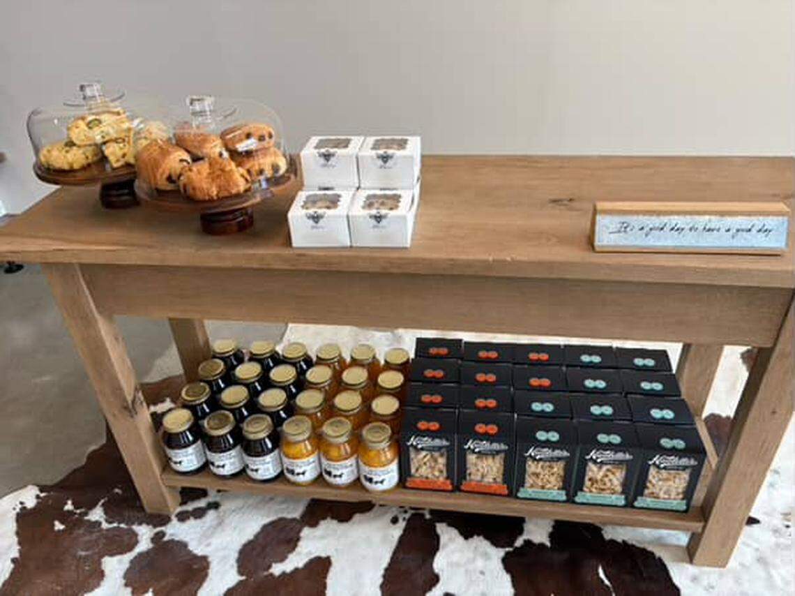 A small wooden counter displays baked goods and various packaged food items. On top of the counter, pastries are under glass domes next to small white boxes of baked goods, and on a shelf underneath, jars of beverages are next to black boxes of snack items.