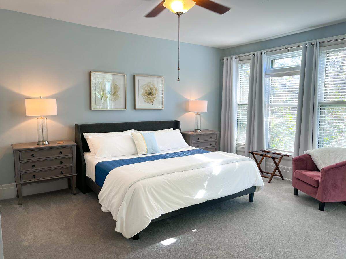 A bright, clean bedroom is shown with a made bed and two nightstands. The room is decorated in a muted color palette with pale blue walls and a dark pink armchair.