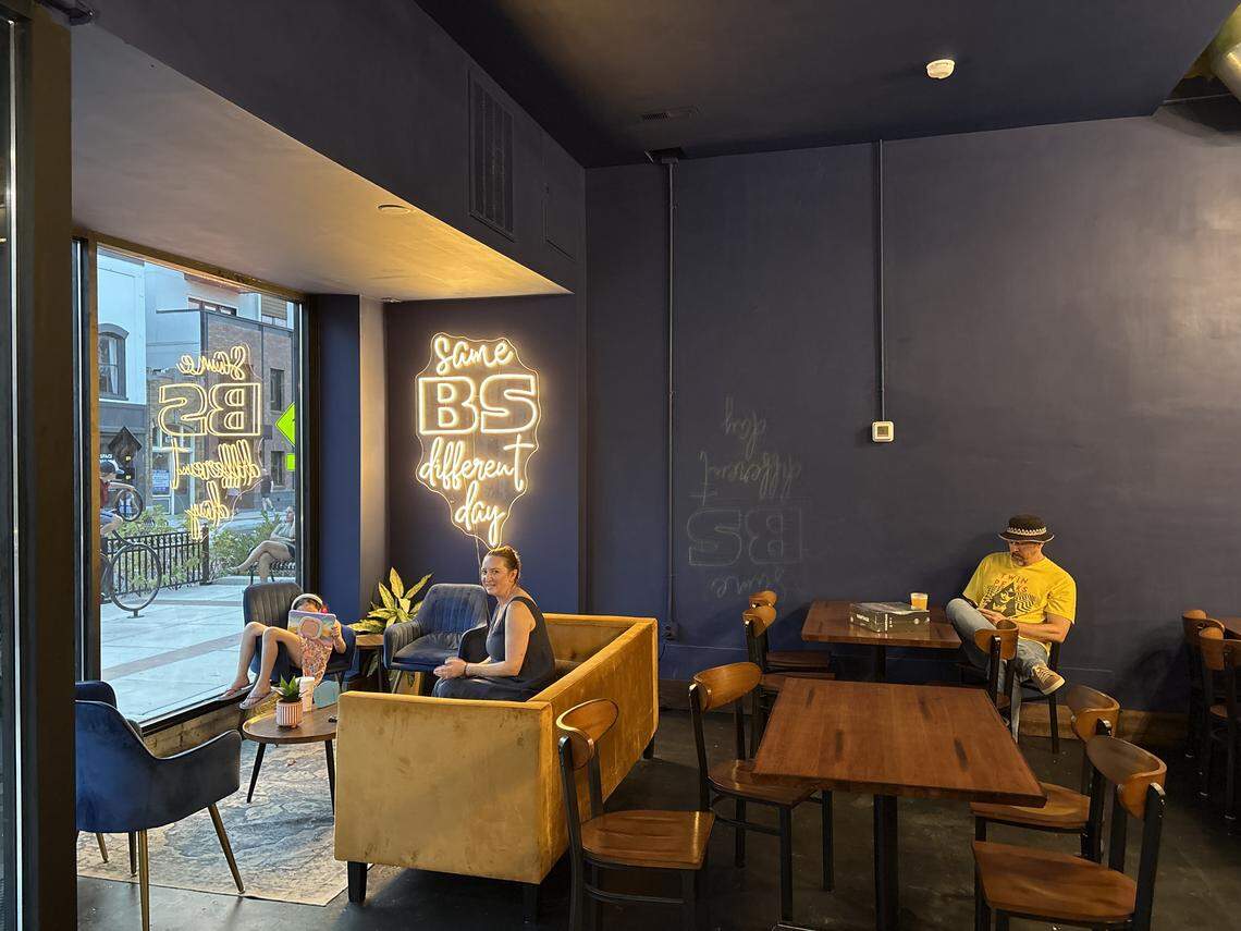 A small cafe with dark blue walls, dark tables, and a yellow couch is occupied by three people. A young girl sits in a chair reading a book, a woman sits smiling on a couch, and a man in a bucket hat sits at a table on his phone.
