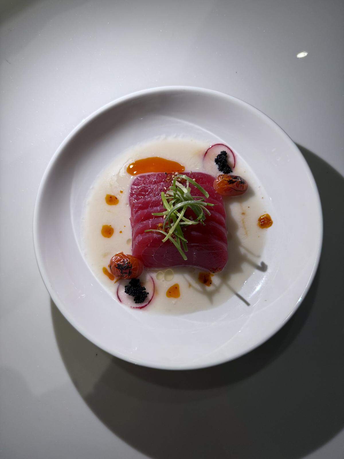 A vibrant, high-angle close-up shot of a restaurant-style dish on a white plate. A large, rectangular block of deep red tuna is the centerpiece, topped with thin green herbs. It rests in a shallow pool of light-colored sauce with drops of reddish oil. Around the tuna are three small, halved red radishes, each topped with a small dollop of black caviar.