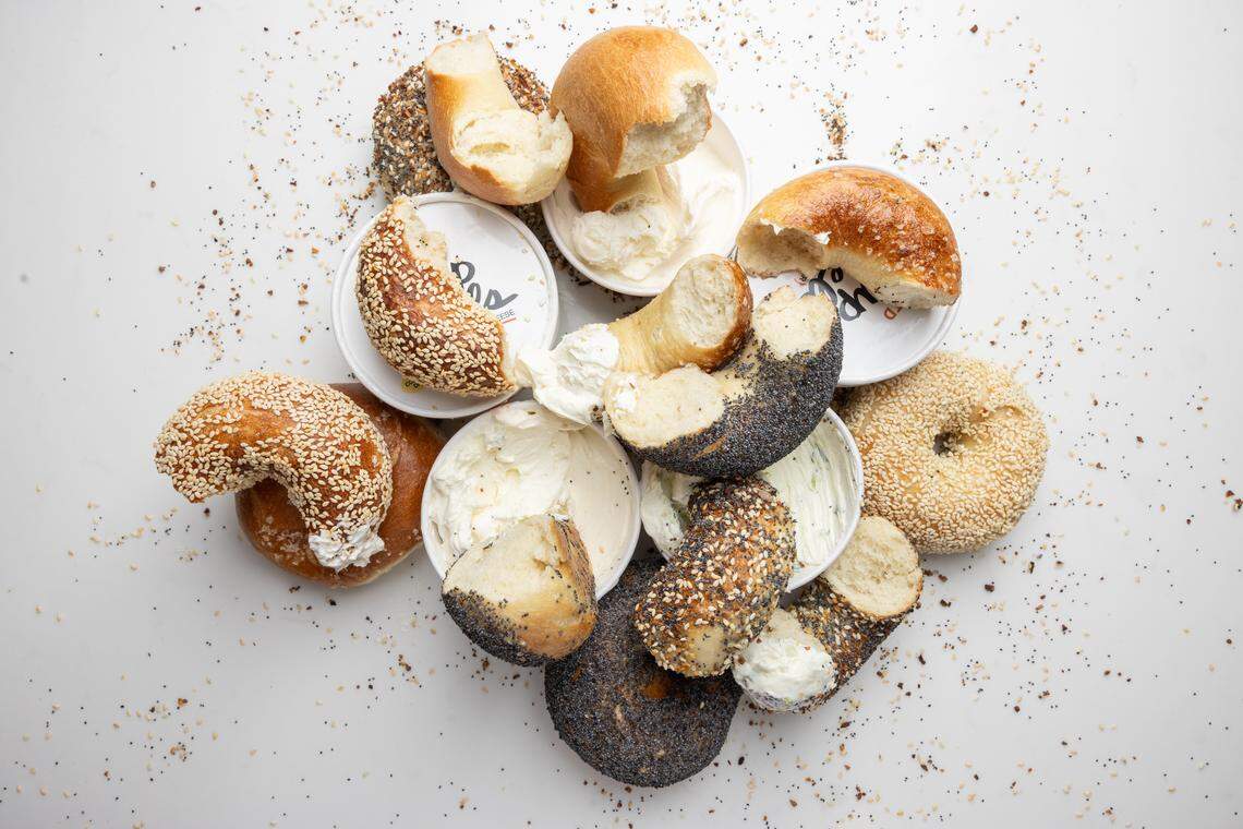 An overhead, artistic lifestyle shot showing several sliced bagels with various toppings, including poppy seed and sesame, scattered on a white surface. The bagel halves are arranged around small white plates holding different types of cream cheese.