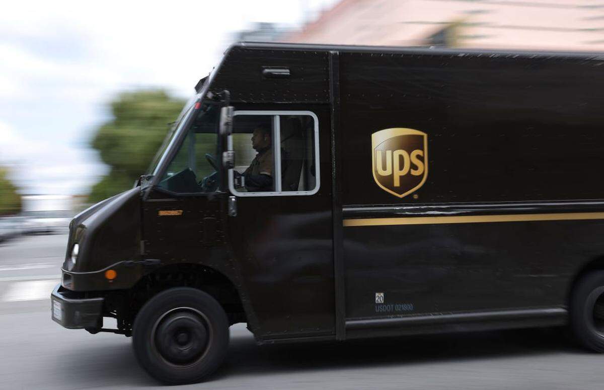 A brown UPS delivery truck drives down a city street, captured in motion blur to indicate speed. A driver in a dark uniform is visible through the passenger-side window looking straight ahead while operating the vehicle.