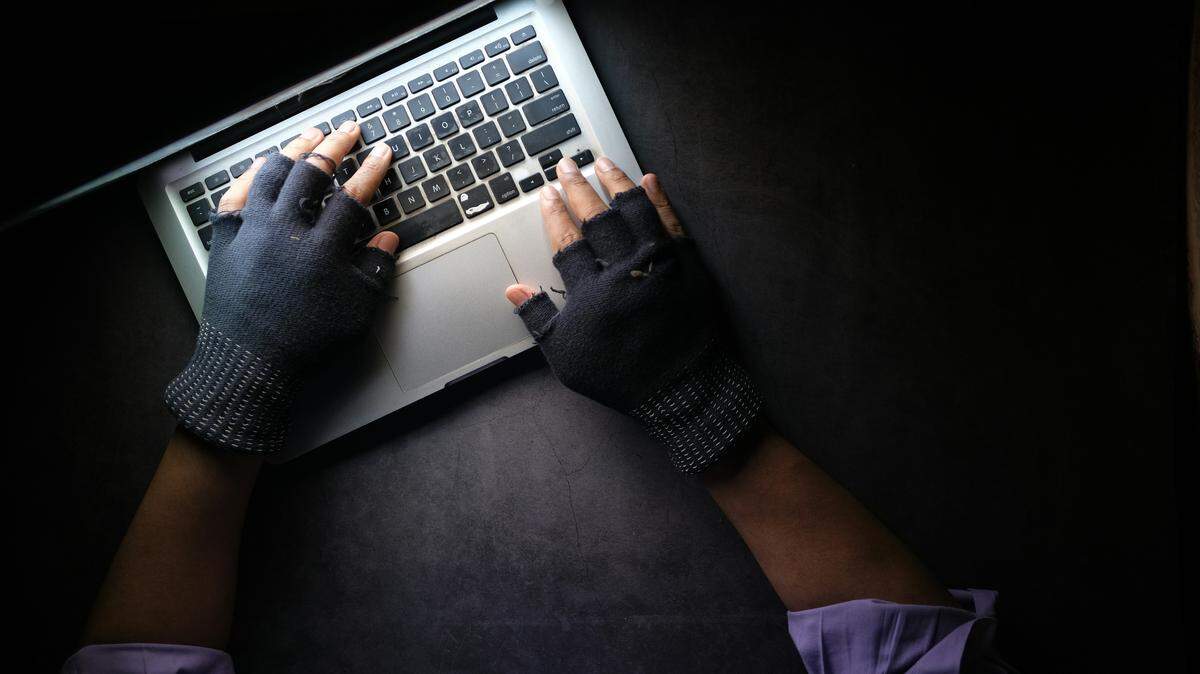 A person wearing dark, fingerless gloves types on a silver laptop keyboard in a dark setting, illuminated primarily by the laptop’s screen. The high angle, overhead shot emphasizes the hands and the keyboard, creating a secretive or focused mood often associated with hacking or intense work.