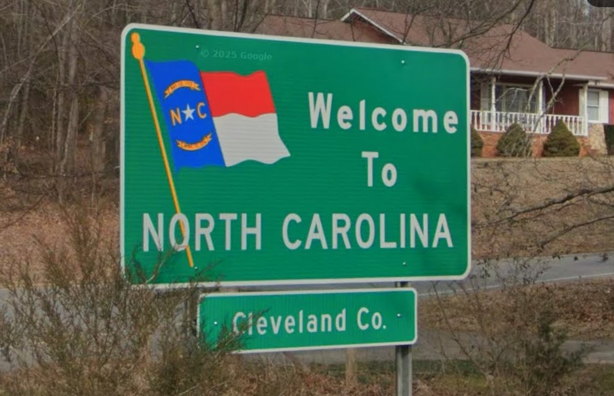 A bright green highway sign welcomes travelers to North Carolina with a stylized image of the state flag waving in the breeze. A smaller sign below it indicates entry into Cleveland County.