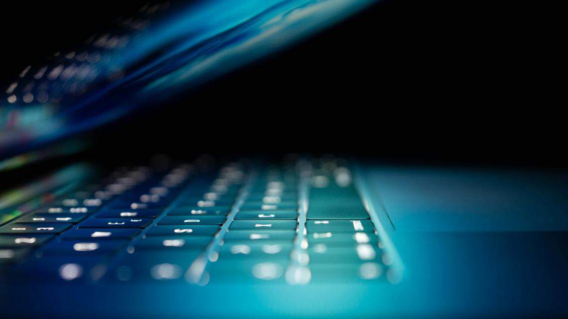 A close-up side view of a laptop keyboard shows the keys glowing with a cool blue and teal light in an otherwise dark room. The focus is on the illuminated keys near the front of the keyboard, suggesting a late-night work session or an image related to cybersecurity or technology.