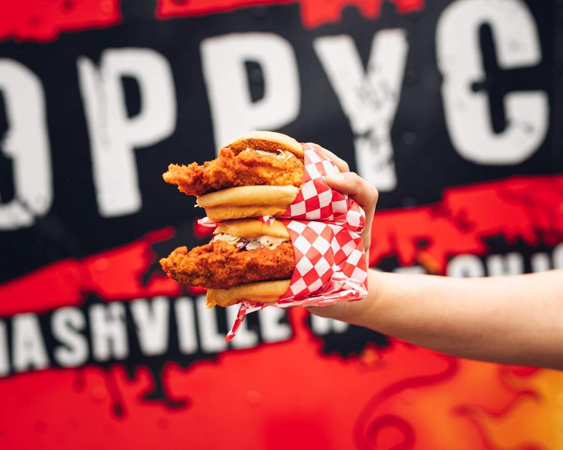 A hand holds up two fried chicken sandwiches wrapped in red and white checkered paper. The background is a red food truck with the words “POPPY’S NASHVILLE HOT CHICKEN” in white.