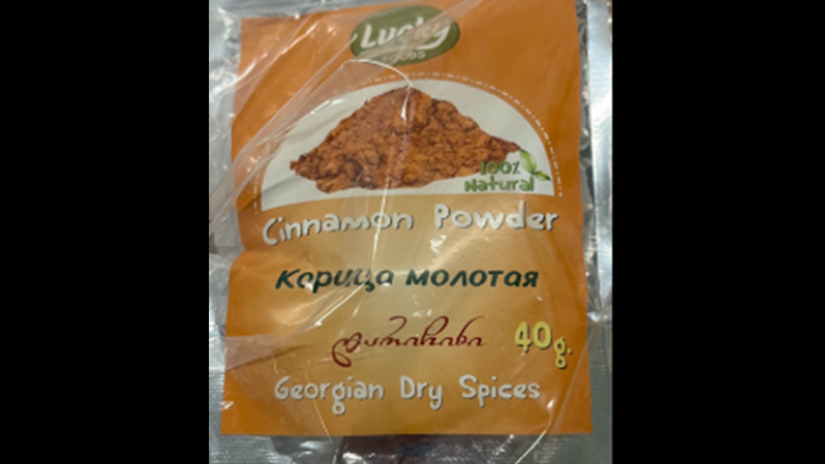 A close-up of a plastic-wrapped package of “Cinnamon Powder” from the “Lucky Foods” brand. The orange label features a small mound of the powder and text in English, Russian (Карица молотая), and Georgian, with a weight of 40g and the label “Georgian Dry Spices.”