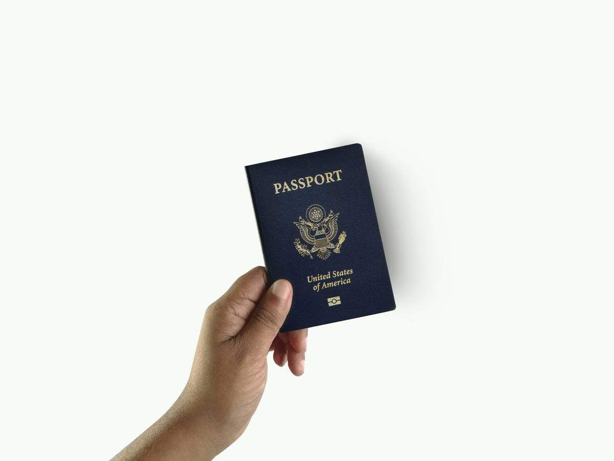 A dark-skinned hand is shown holding a standard U.S. passport, which is navy blue with the national seal embossed in gold. The passport is held slightly at an angle against a stark white background, emphasizing the document’s central role in international travel and citizenship.