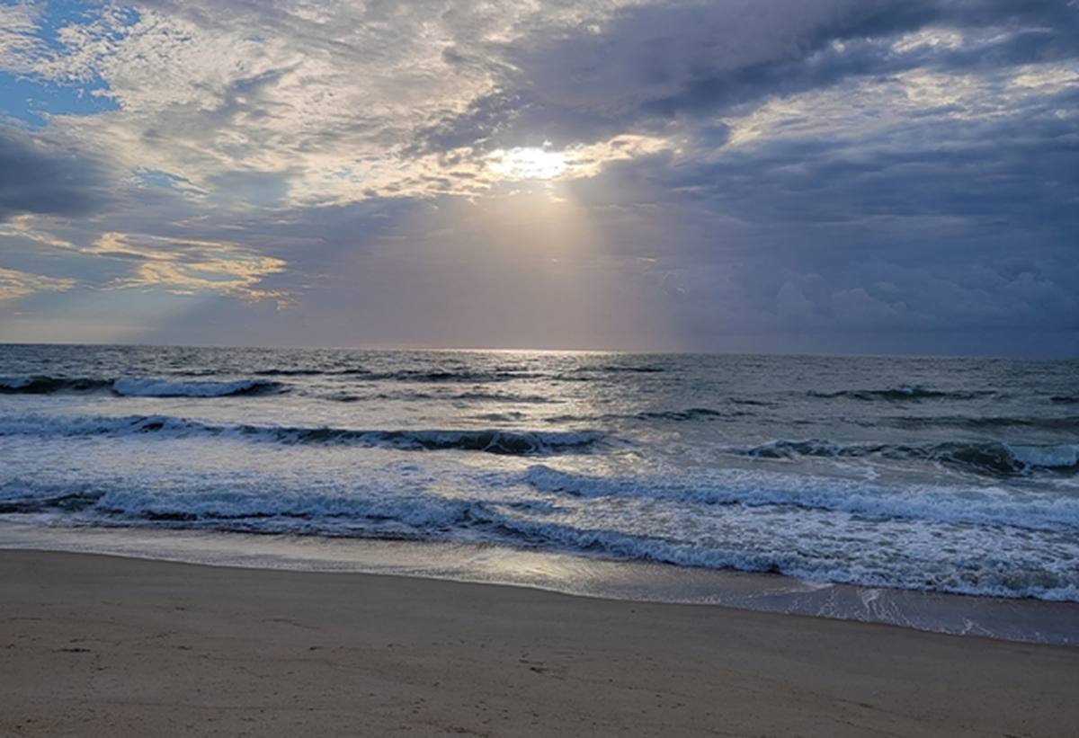 Sunlight breaks dramatically through a thick blanket of dark and white clouds, casting a bright beam across the ocean surface. Gentle waves with white foam roll onto a wide, sandy shoreline in the foreground. The horizon stretches out under a blue sky peeking through the upper left corner of the clouds.