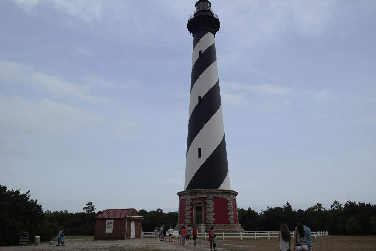 A tall, historic lighthouse featuring distinctive black and white diagonal spirals towers over a grassy field. Tourists wander near a white picket fence at the base of the structure, which includes a small brick outbuilding to the left. The scene is framed by a dense line of trees in the background under an overcast sky.