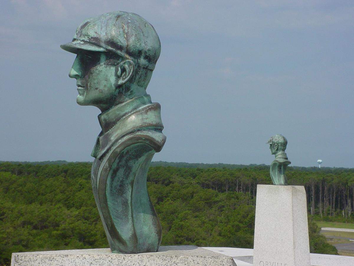 A close-up profile view highlights a green, weathered bronze bust of a man wearing a classic aviator cap. In the background, a second matching bust stands further away on a stone pedestal marked with the name “ORVILLE.” A vast, dense green forest extends toward the horizon under a hazy blue sky.