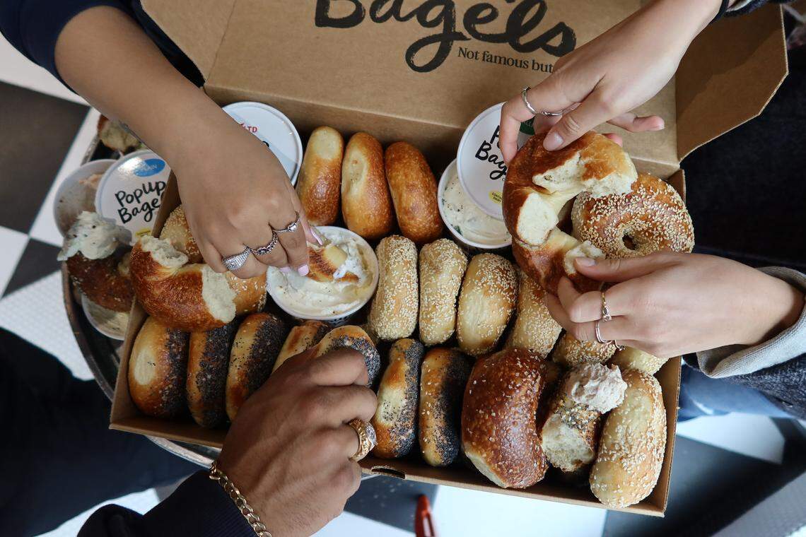 An overhead, artistic lifestyle shot showing several sliced bagels with various toppings, including poppy seed and sesame, scattered on a white surface. The bagel halves are arranged around small white plates holding different types of cream cheese.