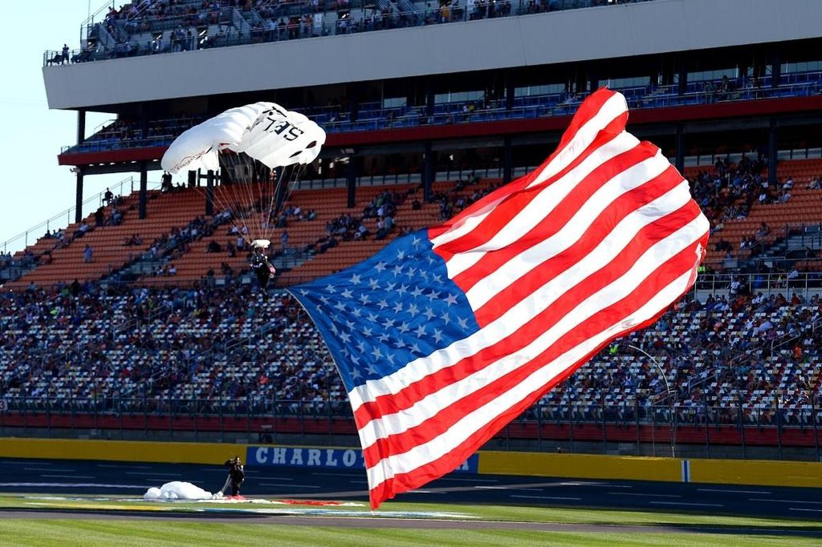 A skydiver carries the American flag onto the infield of Charlotte Motor Speedway before Saturday’s NASCAR qualifying race for the Cup Series all-star race. The race was not completed in time for this edition. Visit ThatsRacin.com for coverage.